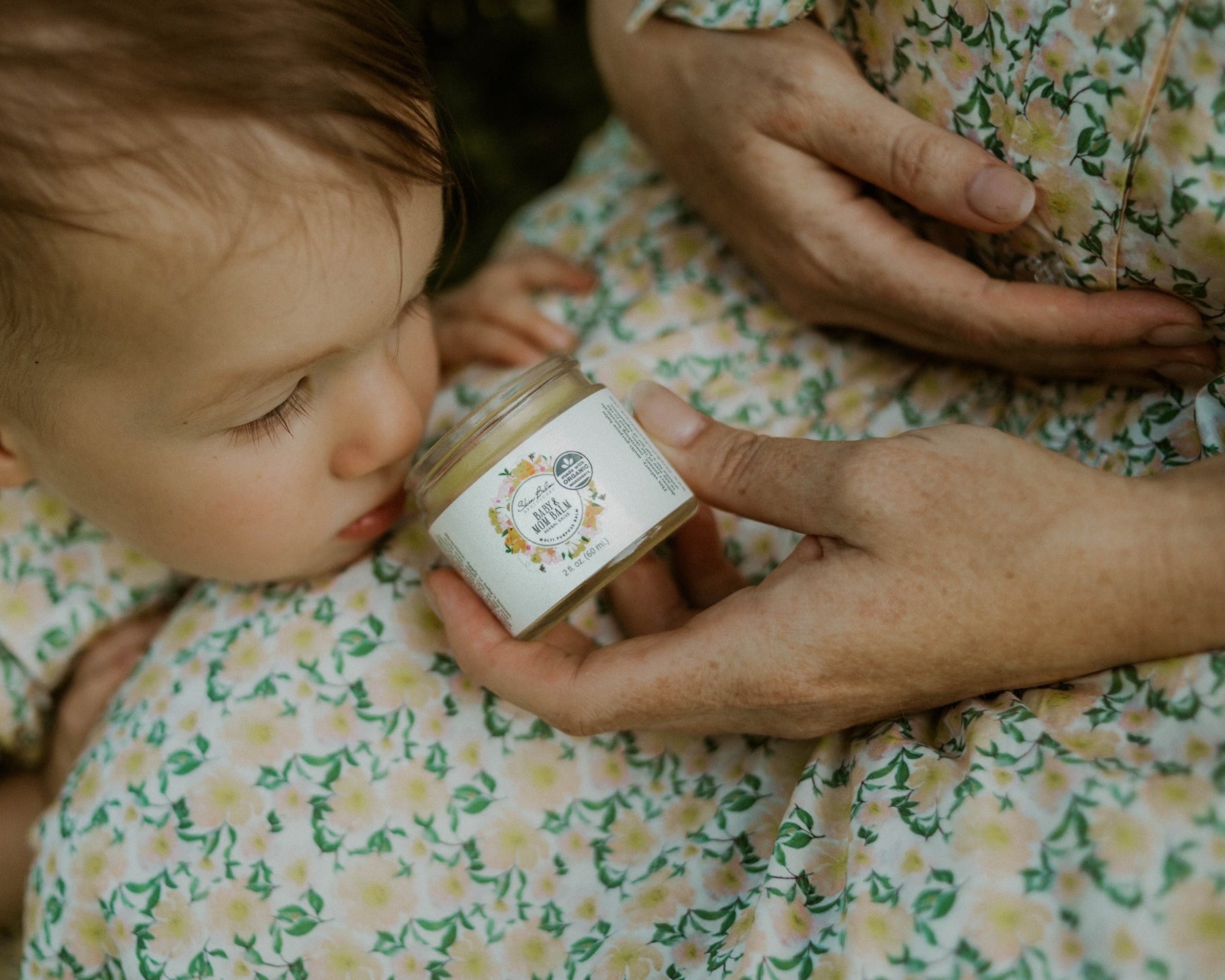 A pregnant woman in a yellow and green floral print dress holds the Baby & Mom Balm in her lap.