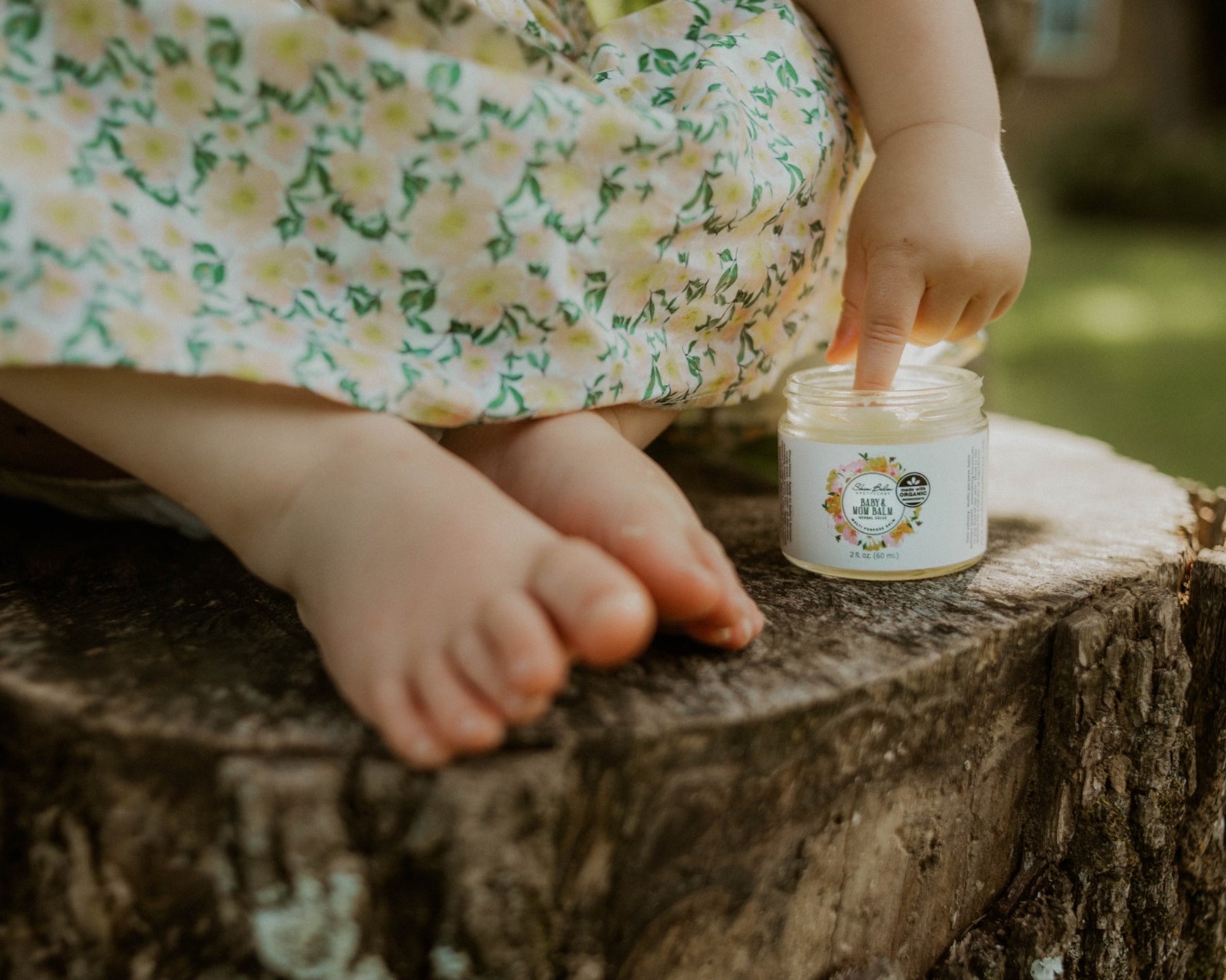 A toddler sits on a tree stump while scooping the Baby & Mom Balm out of the jar with her finger.