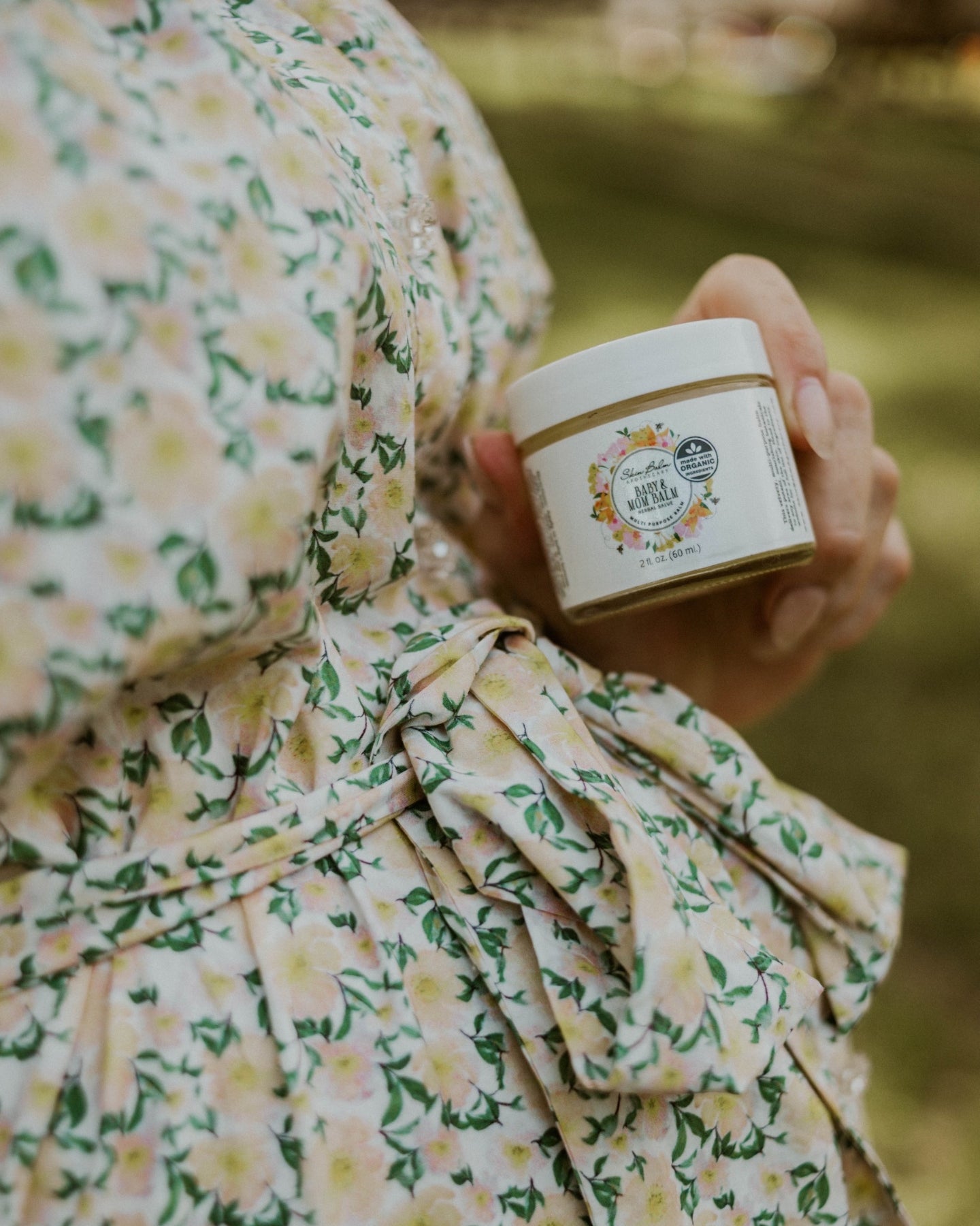 A close-up shot of a pregnant woman in a yellow and green floral print dress holding the Baby & Mom Balm against her bump.