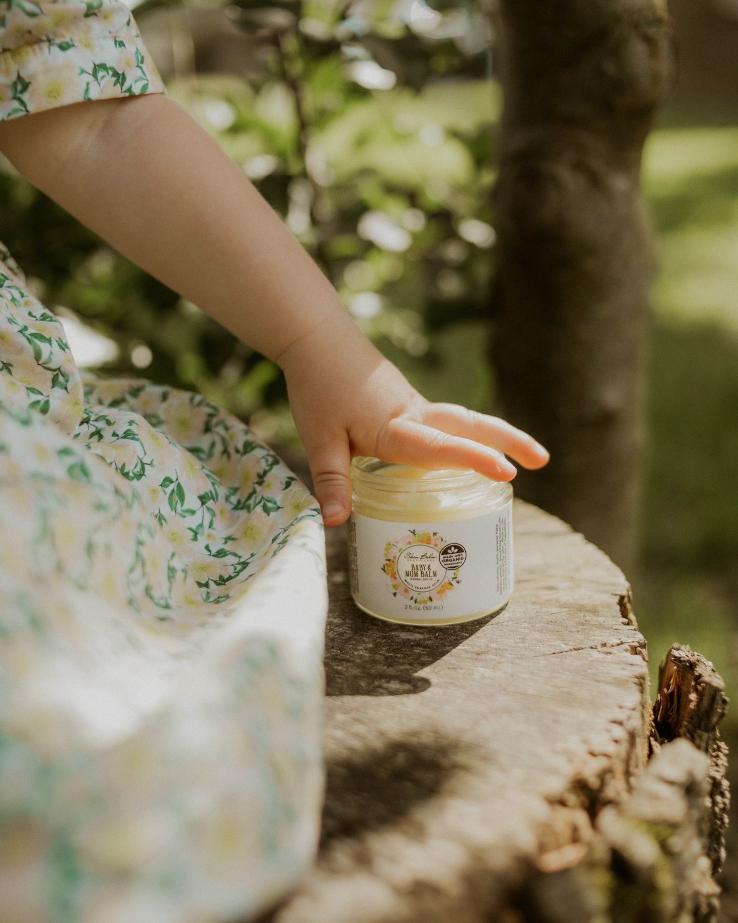 A close-up shot of a toddler resting her hand on the Baby & Mom Balm while sitting on a tree stump.