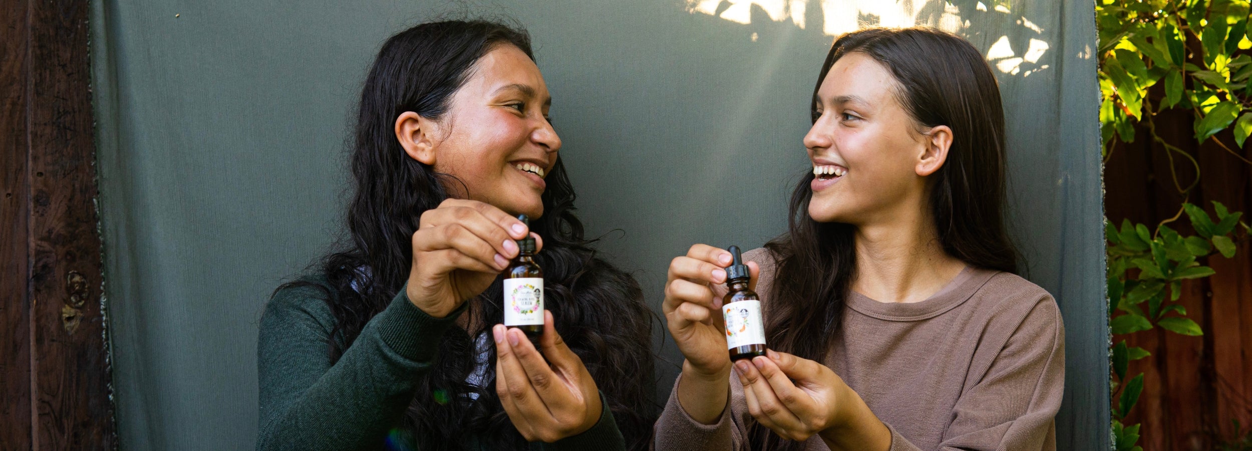 Two women look at each other and smile as they hold up in front of them the Firming Serum and the Purifying Serum.